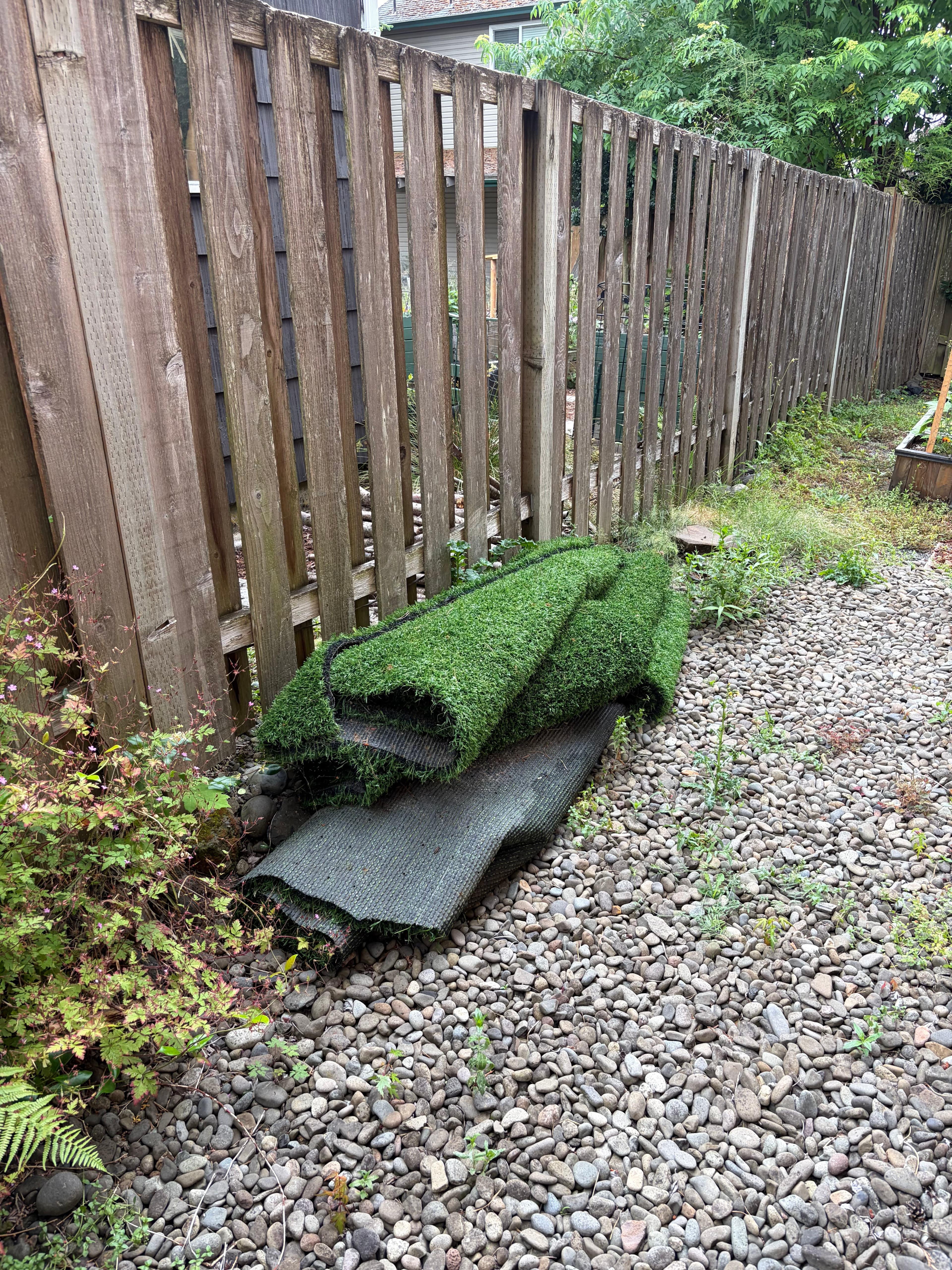 Green artificial turf rolls piled on a pebble ground against a tall wooden fence.